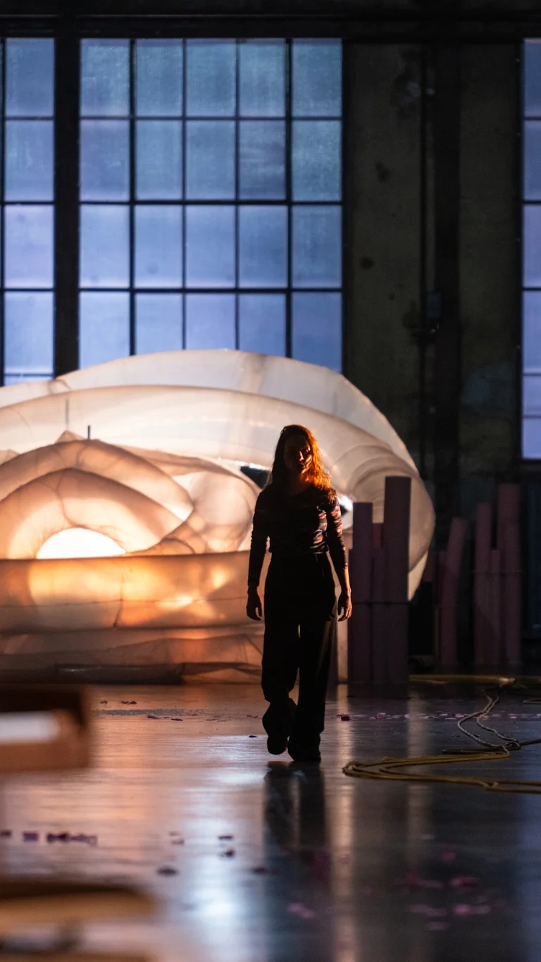 A person walks across a dimly lit stage toward the camera. Behind them, a large white inflatable structure glows in front of tall industrial windows. Cables and scattered pink flower petals lie across the floor.