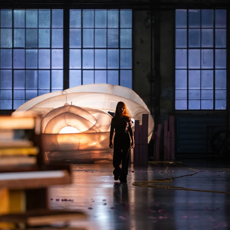 A person walks across a dimly lit stage toward the camera. Behind them, a large white inflatable structure glows in front of tall industrial windows. Cables and scattered pink flower petals lie across the floor.