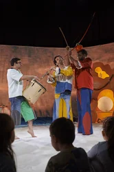 Drei Musiker spielen Flöte, Trommel und ein bogenförmiges Instrument vor Kindern.