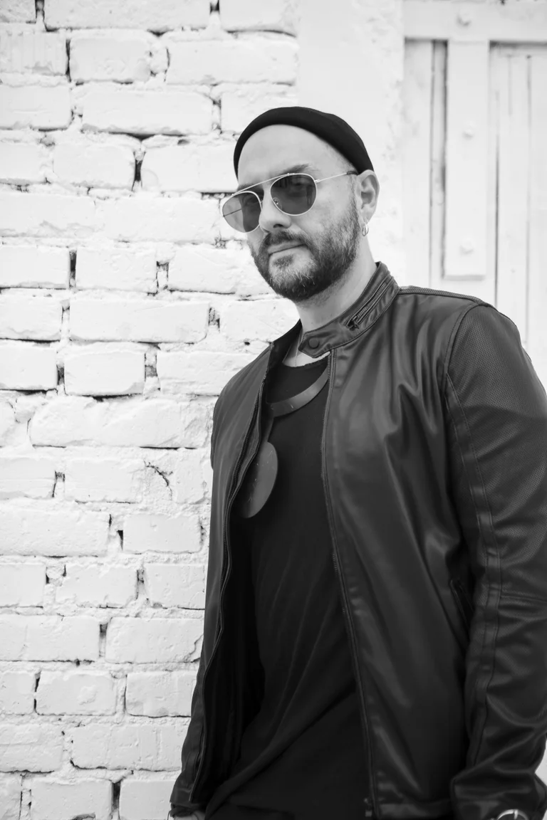 Ein Mann in Sonnenbrille und Lederjacke lehnt an einer Ziegelwand.
