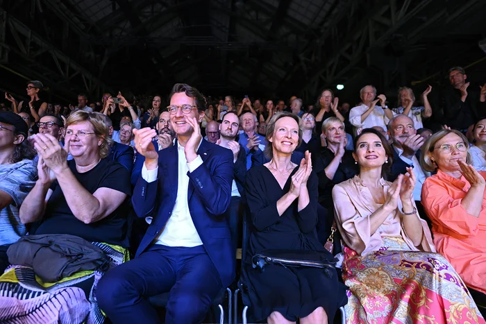 The image shows the opening of the Ruhrtriennale 2024. In the front row, Minister President Hendrik Wüst, Managing Director Dr. Vera Battis-Reese, and State Secretary Gonca Türkeli-Dehnert are seen applauding. They are seated among a packed audience celebrating the event. The atmosphere is festive, marking the start of the significant cultural festival. Everyone appears joyful and in high spirits.