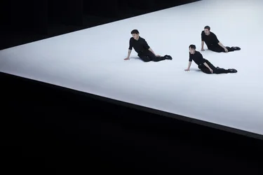 Three performers sit on a slanted, bright stage, supporting themselves with their hands.