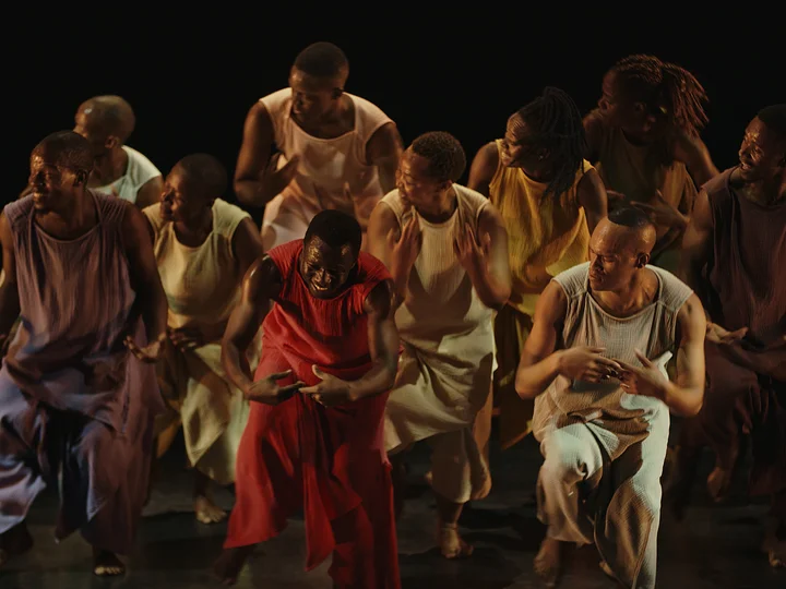 Group of elf dancers in flowing sleeveless costumes in red, yellow and earthy tones performing a dynamic, synchronized choreography on a dark stage.