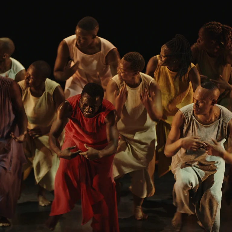 Gruppe von elf Tänzer*innen in fließenden, ärmellosen Kostümen in Rot-, Gelb- und Erdtönen, die auf einer dunklen Bühne synchron eine dynamische Choreografie ausführen.