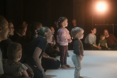Zwei Kleinkinder stehen am Rand einer hellen Bühne und schauen in Richtung des Geschehens. Erwachsene sitzen hinter ihnen auf dem Boden.