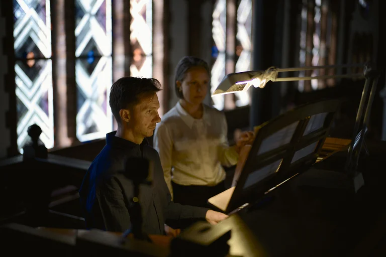James McVinnie and Eliza McCarthy at the organ in the Evangelical Church at the Market in Katernberg during the event 124 Years of Reverb. Both musicians are focused at the organ console, while soft light streams through the large stained-glass windows in the background.