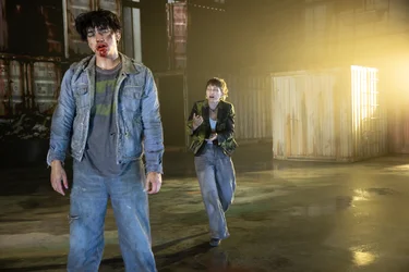Two performers are on a stage with shipping containers. In the foreground, a person dressed in denim stands with their eyes closed and traces of blood on their face. Behind them, another person approaches with outstretched hands. Intense yellow light falls across the stage from the right.