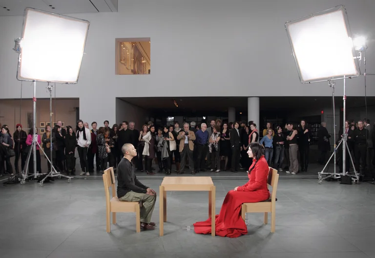 Marina Abramović sits motionless at a table facing a visitor while an audience watches in the museum. The performance “The Artist Is Present” is based on silent encounter and presence.
