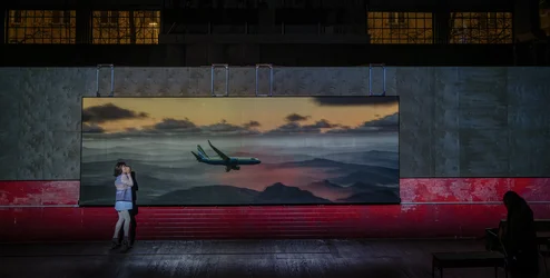 Eine Person steht vor einer großformatigen Videoprojektion eines fliegenden Flugzeugs über einer Landschaft. Eine weitere Person sitzt im Hintergrund auf einer Bank.