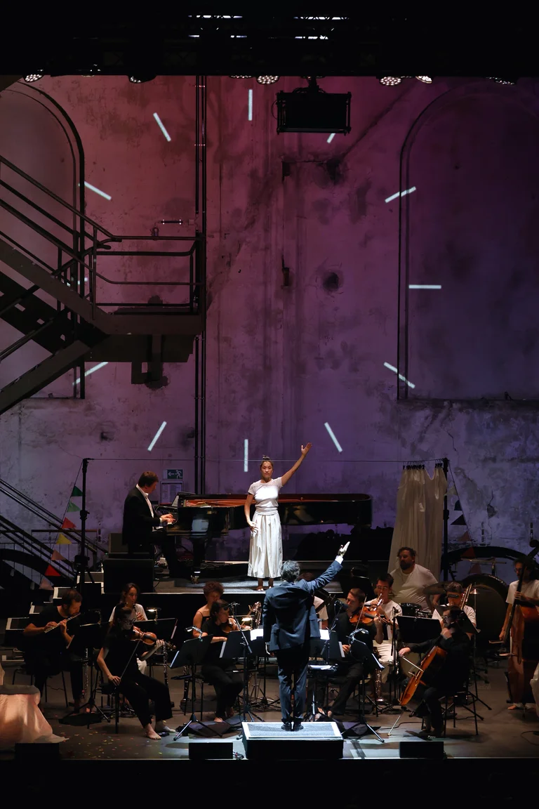The ensemble Les Apaches ! performs on stage in an industrial hall, conducted by Julien Masmondet. Behind them, Sarah Silverblatt-Buser stands with one arm raised in front of a grand piano, where a pianist is playing. White lines of light are projected onto the high wall in the background.