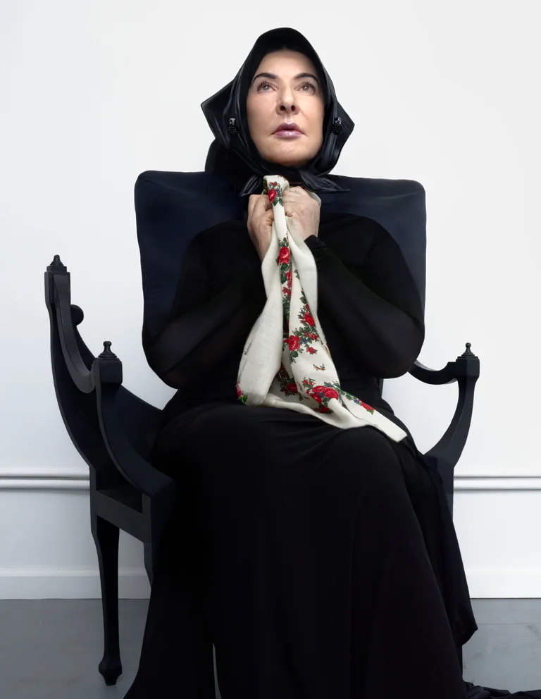 Marina Abramović in black clothing and headscarf sits on a chair, holding an embroidered cloth to her chest.