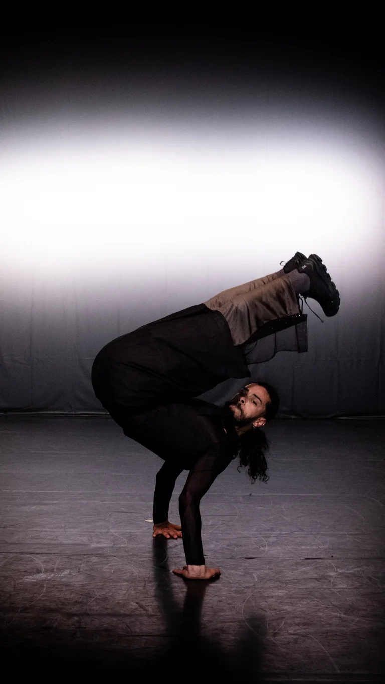 Mohamed Toukabri balances on hands with legs lifted above the head, stage lighting creates a spotlight effect behind.