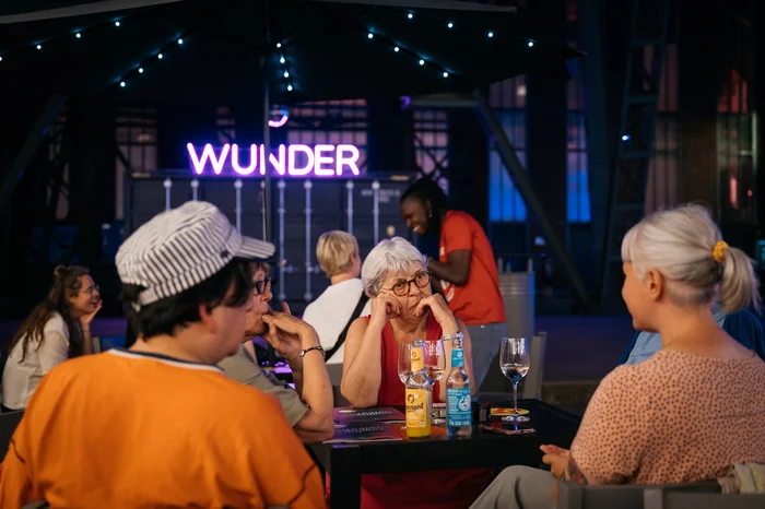 The image shows the festival center Wunderland at the Ruhrtriennale 2024. In the foreground, several people are sitting at a table, casually chatting. Drinks are placed on the table, while the word "WUNDER" glows in neon letters in the background. The atmosphere feels cozy and inviting, with the evening lighting enhancing the special ambiance of the festival center.