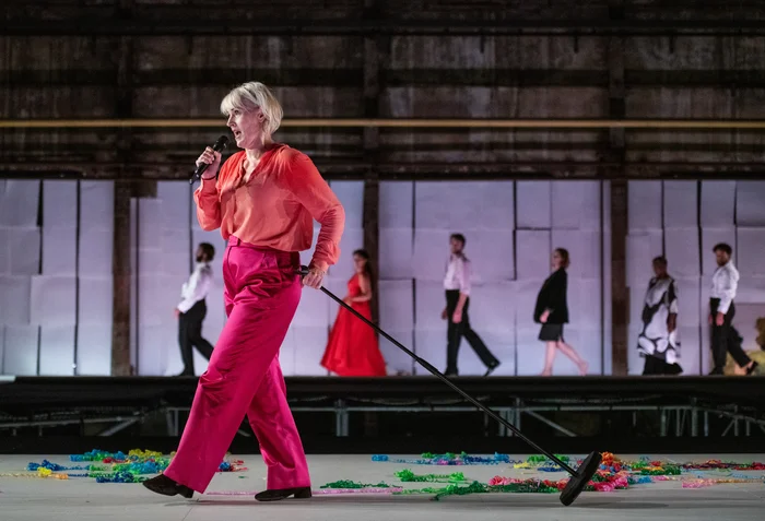 In the foreground, Helena Rasker stands on a brightly lit stage, holding a microphone and dressed in a vibrant pink and red outfit. Colorful paper garlands are scattered around her feet, while several performers walk across an elevated stage in the background. The scene takes place in a former industrial hall.