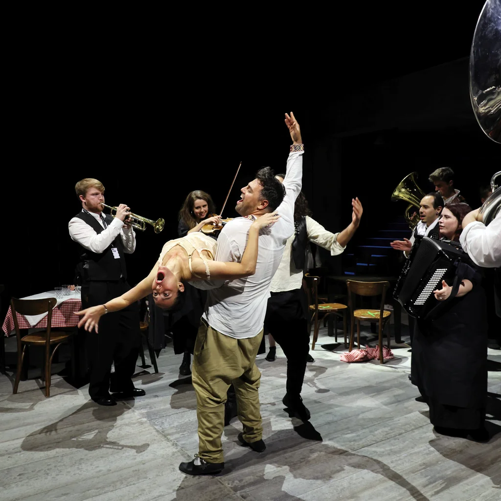 Scene from Balkan Erotic Epic: A person lifts a dancer into a dramatic backbend as a live band with brass and keyboard instruments plays around them. The atmosphere is lively and celebratory.