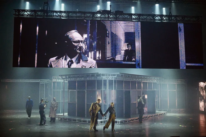 Theatre scene with live video projection: Actors in trench coats and uniforms move around a stage structure made of metal frames, while a large screen above shows close-ups.