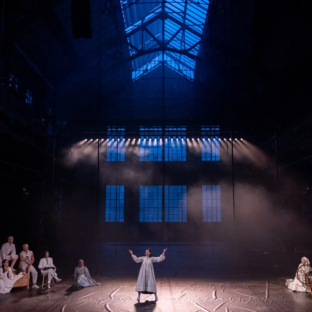 A view into the dark, dimly lit Jahrhunderthalle Bochum. The performers from the 2024 production "Haugtussa"can be seen in the foreground.