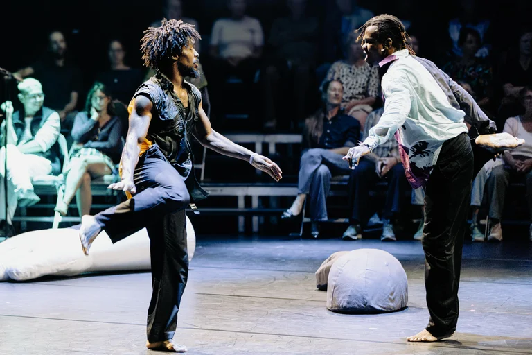 Two dancers stand facing each other, each balancing on one leg. The dancer on the left wears dark clothing. The dancer on the right wears a dirty white shirt. In the background, sacks lie on the stage. Behind them, the audience is seated.