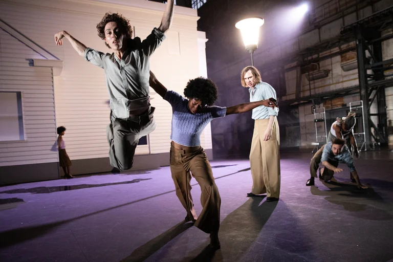 Dancers perform a dynamic choreography on a theater stage. In the foreground, Samuel Planas jumps into the air while Sylvie Sanou moves with outstretched arms. Other performers can be seen in the background, including Lars Eidinger and Larissa Sirah Herden. A white building and a tall streetlamp are visible in the background.
