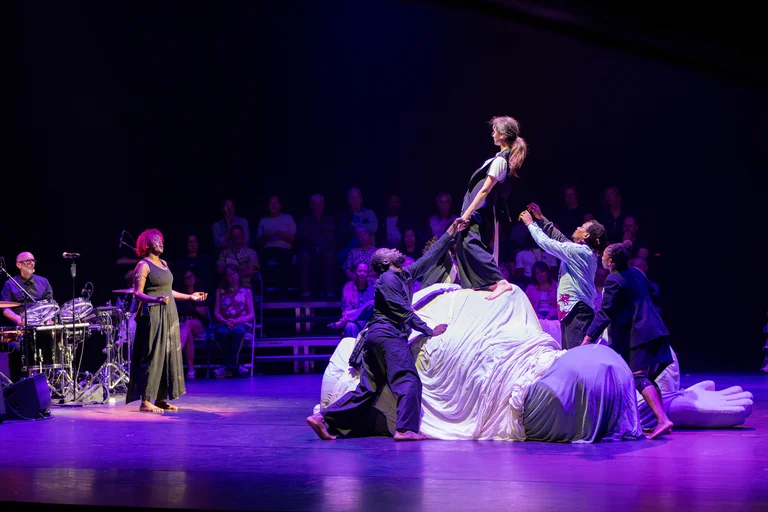 On the left side of the image, a female singer stands on stage in front of a group of four dancers. One female dancer is standing on a structure covered with a white sheet; she is being supported by the others. The audience is seated in the background.