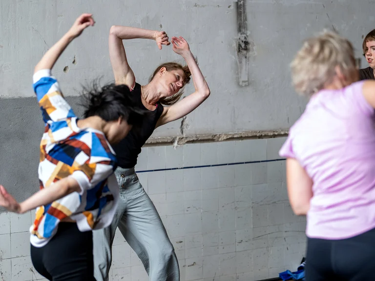 A dancer in colorful clothing swings her upper body backward, surrounded by other dancers.