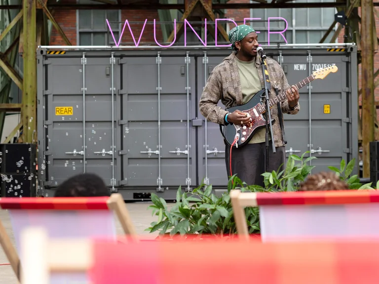 A musician stands in front of a container marked “WUNDER” and plays an electric guitar; deckchairs and plants are visible in the foreground.
