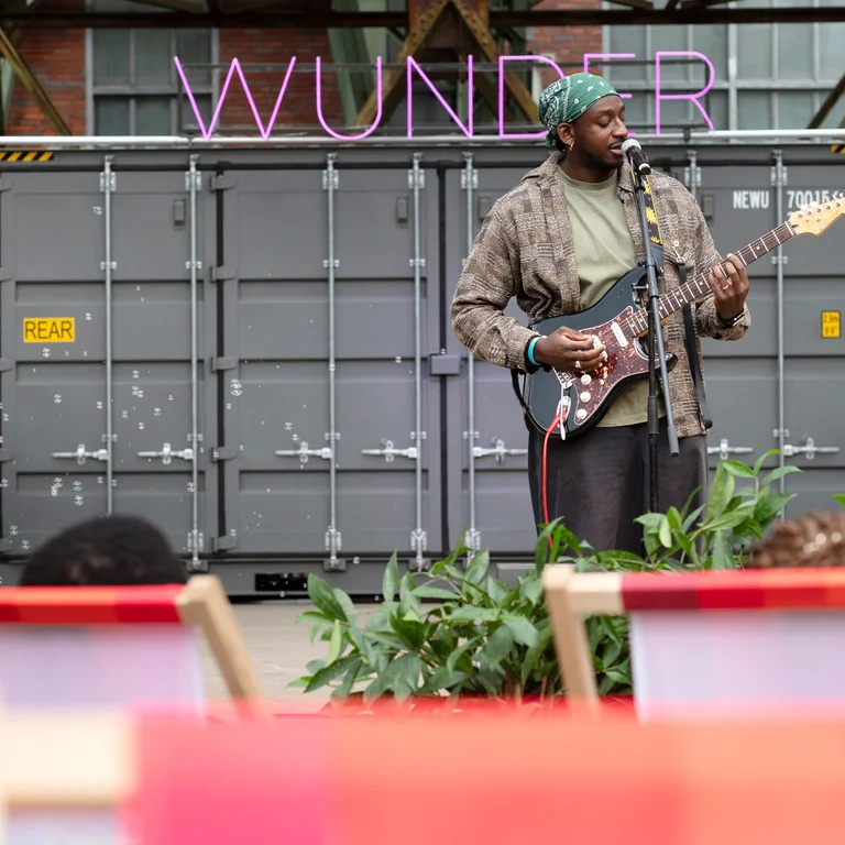 A musician stands in front of a container marked “WUNDER” and plays an electric guitar; deckchairs and plants are visible in the foreground.