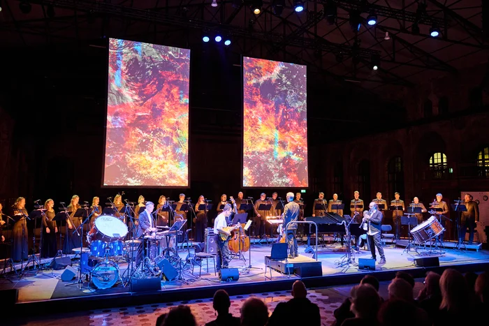 Conductor Florian Helgath with Chorwerk Ruhr and the Bang on a Can All-Stars on stage, with two large projections of abstract red and blue patterns in the background.