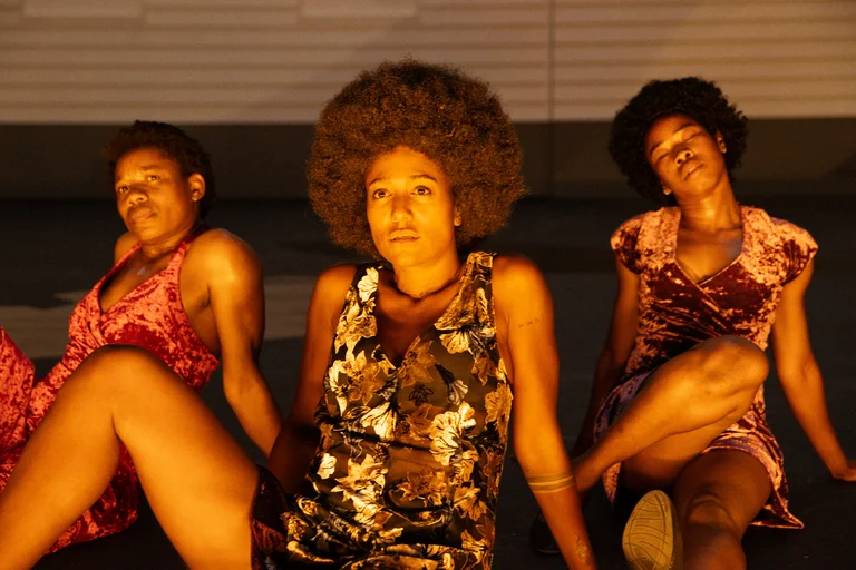 Ida Faho, Larissa Sirah Herden, and Sylvie Sanou are seated on a theatre stage in front of a light-colored house facade. They wear colorful dresses made of shimmering fabric and have Afro hairstyles. Bathed in warm orange light, they gaze in different directions with intense, expressive faces.
