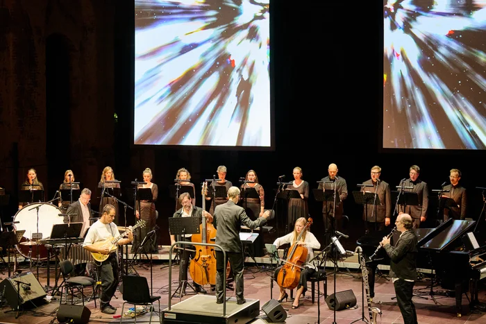 Conductor Florian Helgath with Chorwerk Ruhr and the Bang on a Can All-Stars on stage, with projections of abstract light beams in the background.