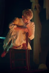 Joyce DiDonato in heller Kleidung sitzt barfuß auf einem Stuhl, den Körper eng zusammengezogen, den Kopf nachdenklich auf die Hand gestützt; warmes Bühnenlicht hebt die Szene vor dunklem Hintergrund hervor.