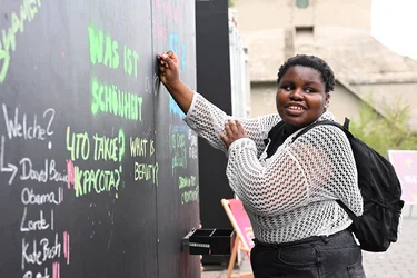 A young woman writes with colorful chalk on a board that is asking "What is beauty?" She smiles at the camera.
