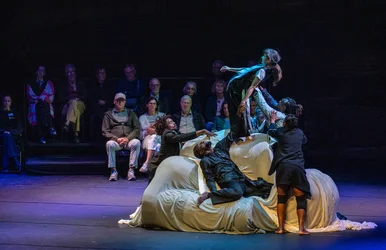 Several performers dressed in dark clothing move together on and around a large object draped in light-colored fabric. One performer is supported and lifted by the others as the audience watches from close by.
