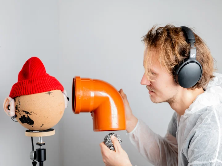 A person wearing headphones holds a large orange pipe piece in front of a globe wearing a red beanie. The scene appears experimental, focusing on acoustic perception.