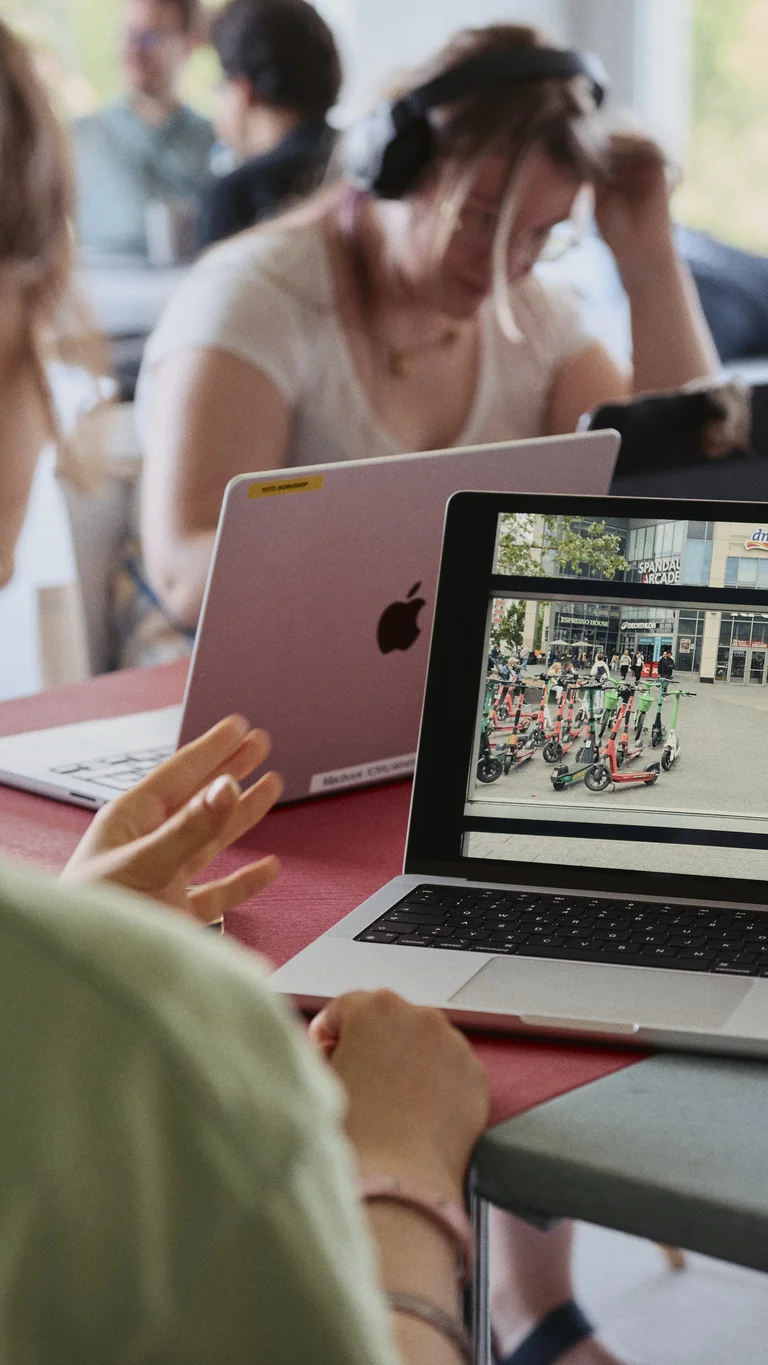 Mehrere Personen sitzen an einem Tisch und arbeiten gemeinsam an digitalen Projekten. Im Vordergrund betrachtet eine Person einen Laptop, auf dessen Bildschirm ein Foto einer städtischen Szene zu sehen ist, und erklärt etwas mit einer Handbewegung. Weitere Teilnehmende arbeiten im Hintergrund an Laptops oder hören zu.
