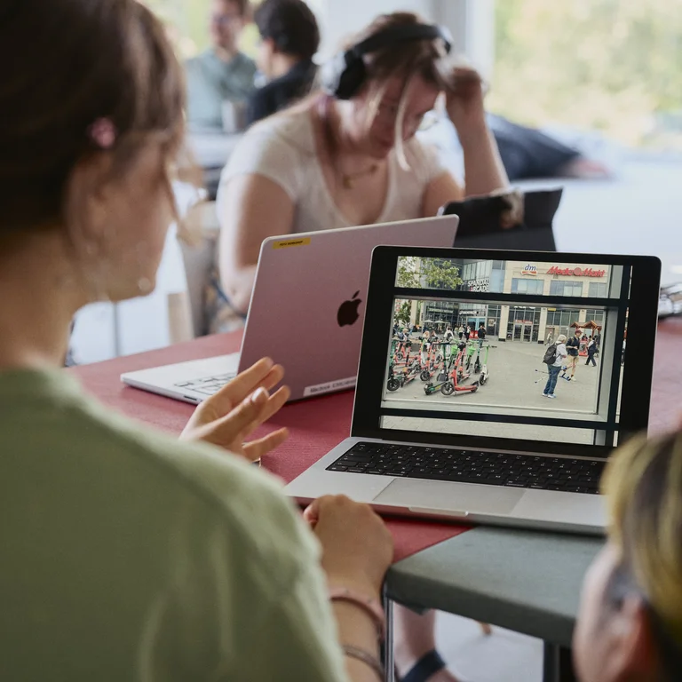 Mehrere Personen sitzen an einem Tisch und arbeiten gemeinsam an digitalen Projekten. Im Vordergrund betrachtet eine Person einen Laptop, auf dessen Bildschirm ein Foto einer städtischen Szene zu sehen ist, und erklärt etwas mit einer Handbewegung. Weitere Teilnehmende arbeiten im Hintergrund an Laptops oder hören zu.