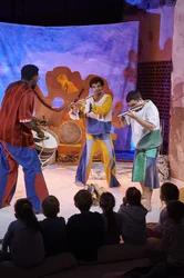 Drei Musiker spielen Trommel und Flöten vor einem jungen Publikum.