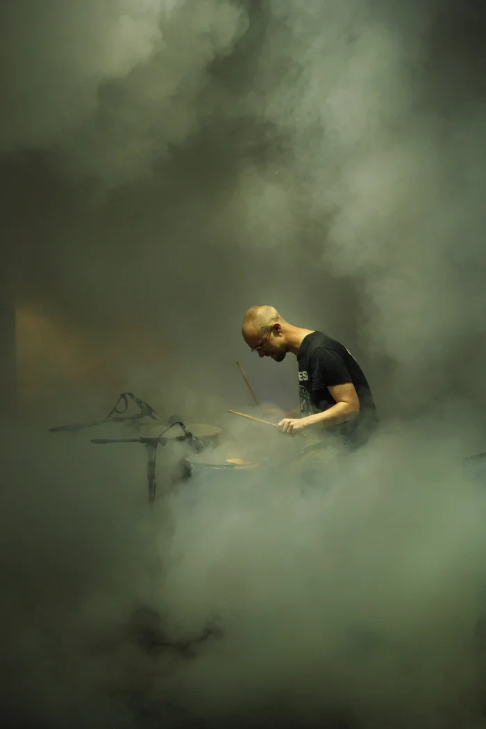 Sam Slater plays the drums, surrounded by dense clouds of fog. Only he and parts of the drum set are visible in the diffuse light.