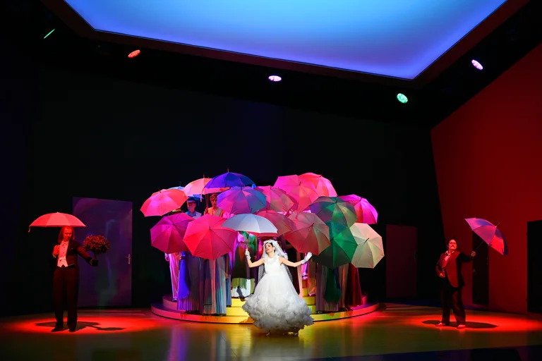 A group of people stand on a stage and form a circle with colourful umbrellas.