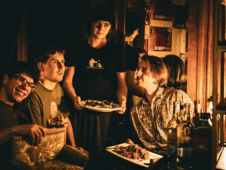 The four band members of Favourite Sidewalk Couch sit and stand around a dining table in warm light, talking in a cosy, home-like interior.