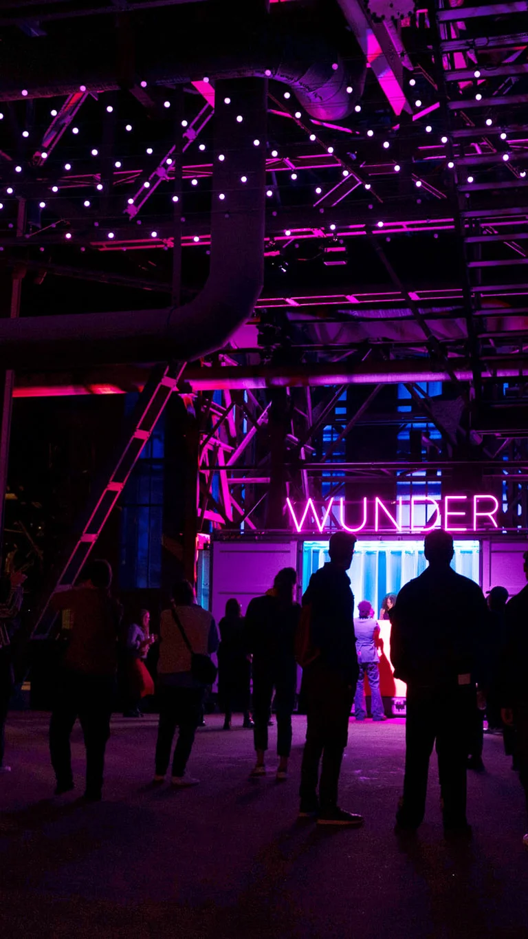 People gather beneath an illuminated steel structure bathed in pink-purple light; the word “WUNDER” glows in the background.