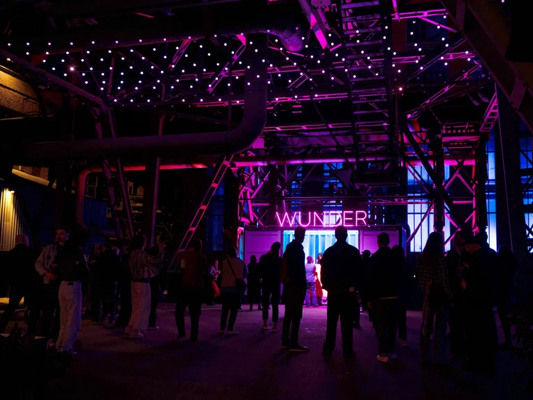 People gather beneath an illuminated steel structure bathed in pink-purple light; the word “WUNDER” glows in the background.
