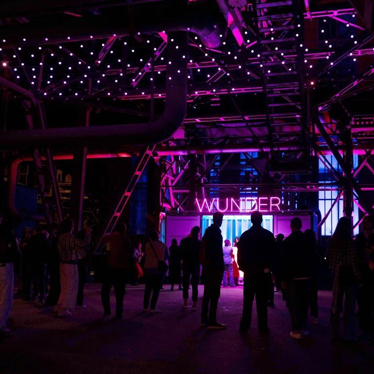People gather beneath an illuminated steel structure bathed in pink-purple light; the word “WUNDER” glows in the background.