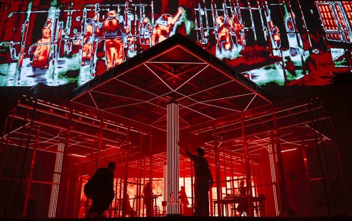 Großprojektion farbverzerrter Silhouetten über einer rot erleuchteten Bühnenszene.