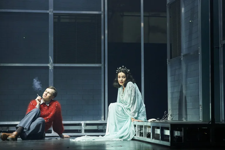 Two actors sit on stage, one in a red sweater smoking, the other in a light blue costume with a star crown.