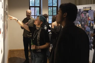 Besucher:innen betrachten Fotografien an einer Ausstellungswand. Eine Person zeigt auf ein Bild.