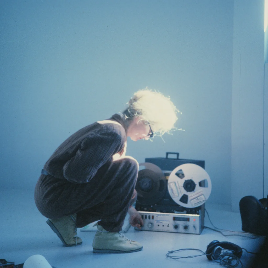Eine Person in Arbeitskleidung kauert in einem minimalistisch eingerichteten, hellblau ausgeleuchteten Raum neben einem offenen Tonbandgerät und bedient dessen Regler; auf dem Boden liegen Kopfhörer und Kabel. Das Licht fällt intensiv auf ihr Haar.