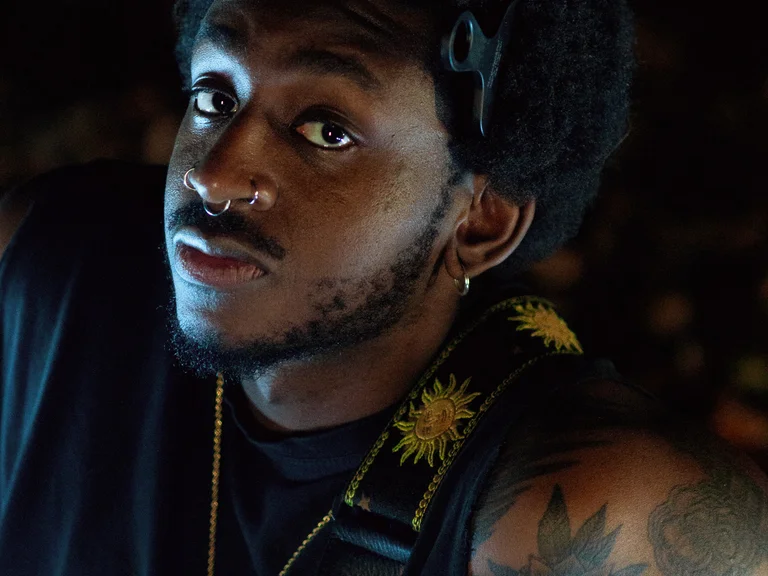 Portrait of Joel Joao with guitar in front of a dark background.