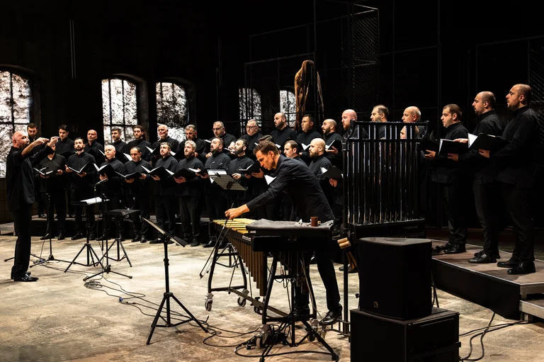 A male choir sings in a hall, in front of them the conductor and a person on the vibraphone.
