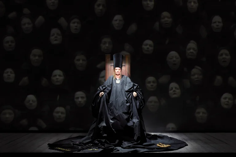In a spotlight, a person sits on a chair wearing a floor-length black robe and tall hat; many faint faces float in the dark background.